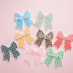 May include: A collection of personalized bow-shaped ornaments in various colors and patterns, including gingham and leopard print. Each bow features a name in a cursive font, with a scalloped edge. The background is a solid pink.