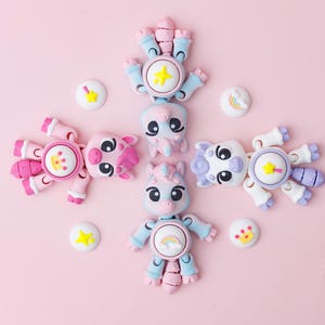May include: A collection of colorful, articulated animal figures, including a pig, octopus, unicorn, and cow, arranged on a pink background. Each figure has a circular design with a star or rainbow. Small, round, decorative pieces with star and rainbow designs are also present.