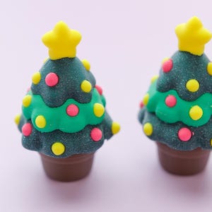 May include: Two miniature Christmas tree decorations. Each tree is dark green with colorful pink and yellow dot ornaments, a bright yellow star on top, and a brown pot base. The trees are set against a soft pink background.