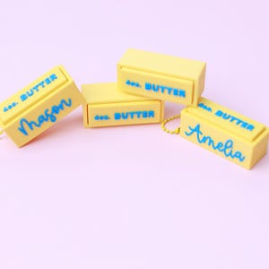 May include: Four yellow rectangular butter-shaped keychains with blue text. Each features the embossed text 4oz. BUTTER. Two keychains are personalized with the blue script names Mason and Amelia, and two include yellow ball chains.