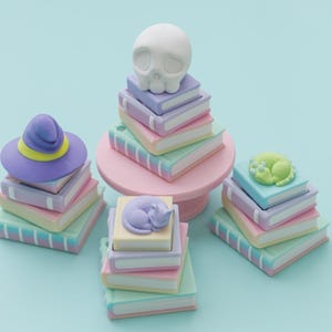 May include: A collection of whimsical miniature book stacks in pastel colors, topped with a witch hat, a skull, a sleeping cat, and a sleeping frog. The books are in shades of pink, blue, and green, with white spines. The background is a light blue.