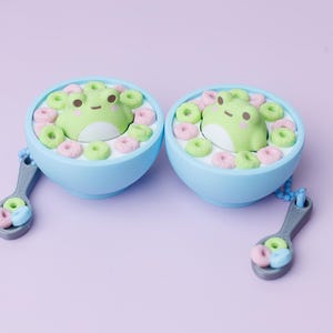 May include: Two light blue bowls filled with white milk and small green and pink ring-shaped cereal pieces. A small green frog figure sits in the center of each bowl. Each bowl is attached to a grey spoon charm containing more cereal pieces.