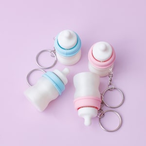 May include: Four baby bottle keychains in white, blue, and pink. The keychains have silver rings and are arranged on a light purple background. The bottles have a classic design, with a nipple and a ring around the middle.