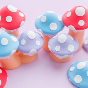 May include: A collection of colorful mushroom figurines in red, blue, and purple with white polka dots. The mushroom caps are rounded, and the stems are a light orange color. The background is a soft pink.