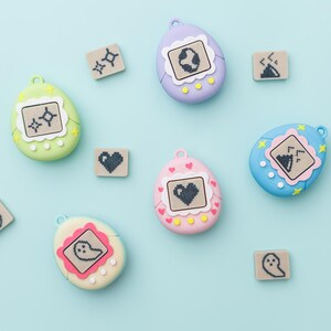 May include: A collection of colorful, egg-shaped digital pet toys in shades of green, purple, pink, and blue, each with a small screen displaying pixelated graphics. Small square blocks with pixelated icons are scattered around the toys.