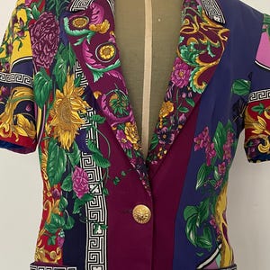 May include: A short-sleeved blazer with a vibrant floral and geometric pattern. The jacket features a mix of colors including purple, blue, yellow, and red, with a gold button closure and pocket details. The design includes sunflower and floral motifs.