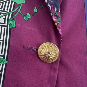 May include: Close-up of a purple blazer with a gold button. The button features a sun design. The blazer has a floral and geometric patterned silk lining in green, white, and purple.