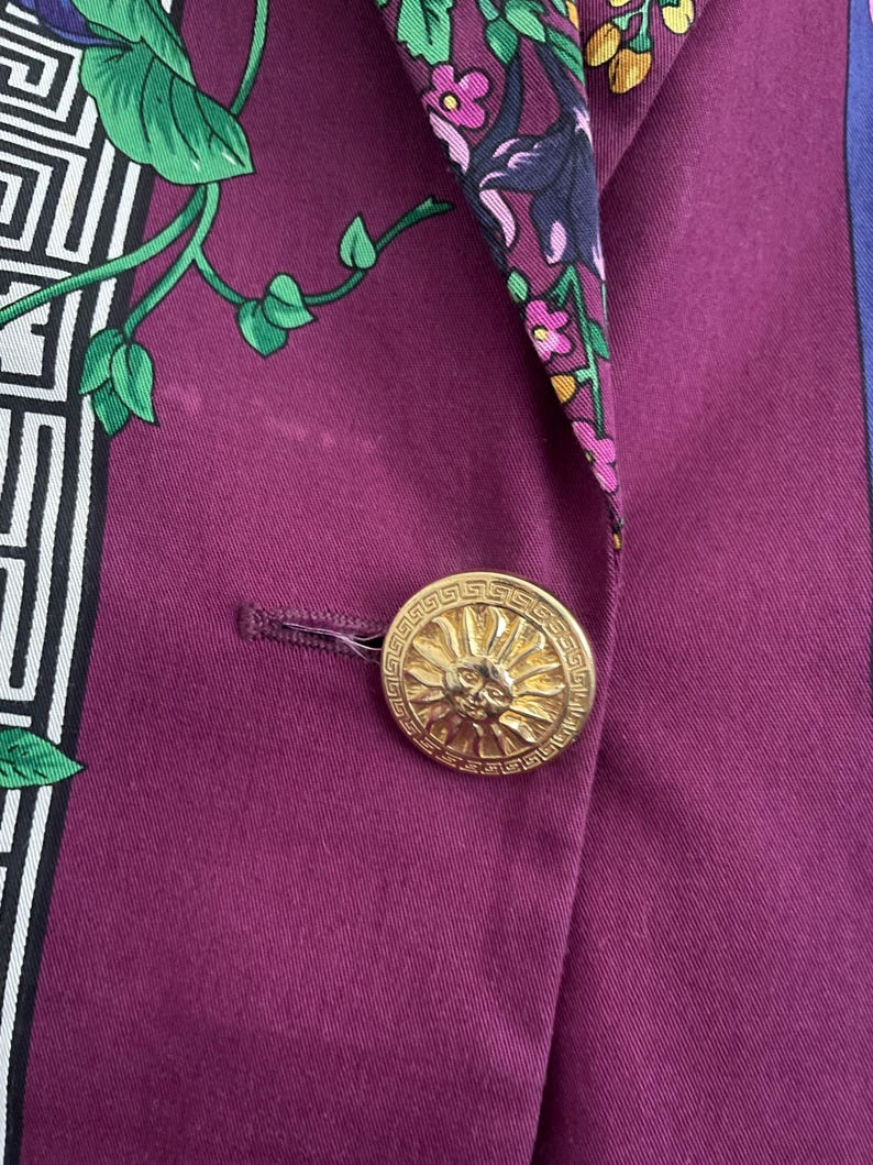 May include: Close-up of a purple blazer with a gold button. The button features a sun design. The blazer has a floral and geometric patterned silk lining in green, white, and purple.