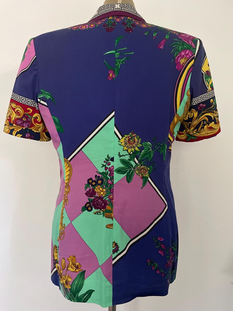 May include: A short-sleeved blazer with a vibrant, patterned design. The blazer features a color block design with purple, green, and blue panels. Floral and gold accents adorn the sleeves and collar, creating a bold, eye-catching look.