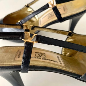 以下が含まれることがあります： ゴールドのアクセントと「miss V valentino garavani」と書かれたゴールドのラベルが付いた、黒い革のハイヒールシューズです。
