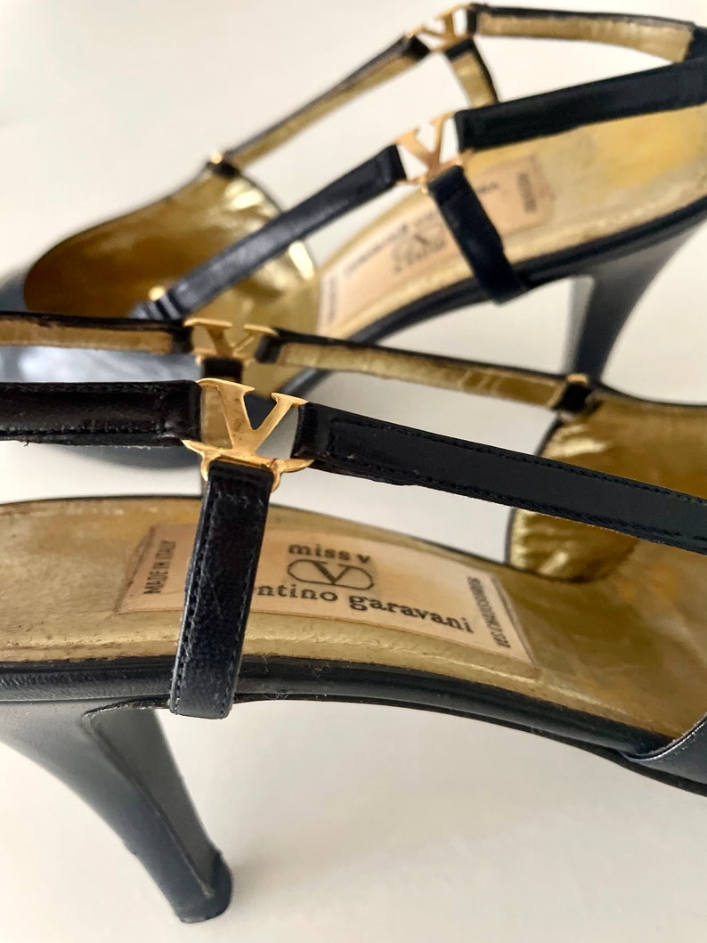 以下が含まれることがあります： ゴールドのアクセントと「miss V valentino garavani」と書かれたゴールドのラベルが付いた、黒い革のハイヒールシューズです。