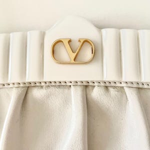 Pode incluir: Uma bolsa de m&atilde;o de couro branco com um logotipo V de metal dourado na frente.
