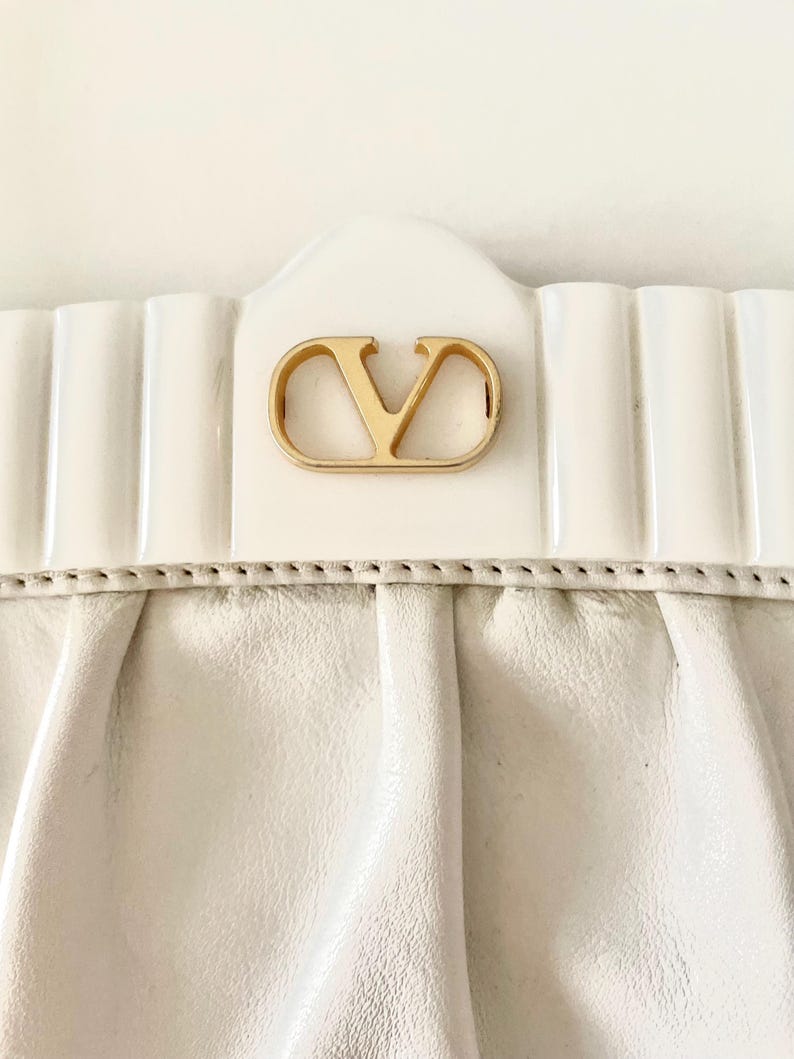 Pode incluir: Uma bolsa de m&atilde;o de couro branco com um logotipo V de metal dourado na frente.