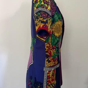 May include: A short-sleeved, vintage blazer in shades of blue, purple, red, and yellow. The blazer features a floral and geometric pattern, with gold-colored buttons. The design includes sunflowers and a Greek key border.