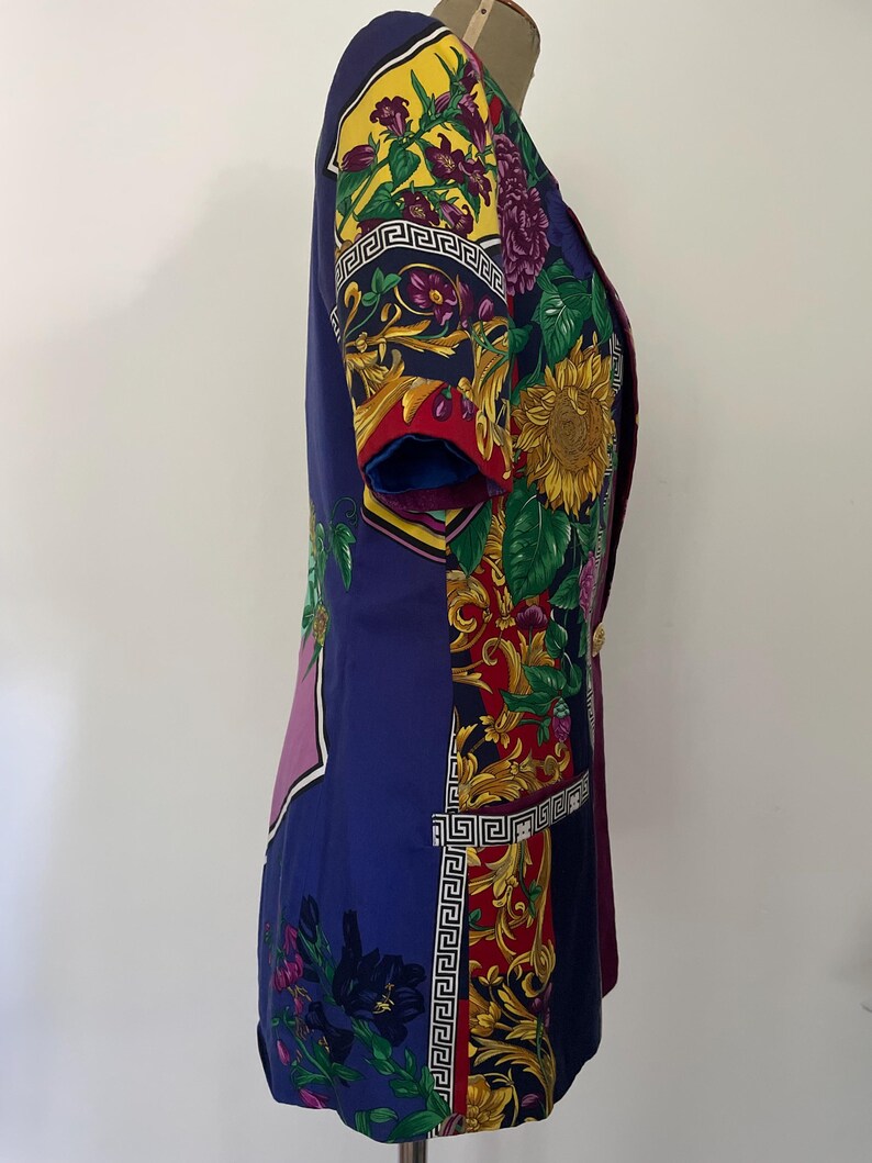May include: A short-sleeved, vintage blazer in shades of blue, purple, red, and yellow. The blazer features a floral and geometric pattern, with gold-colored buttons. The design includes sunflowers and a Greek key border.