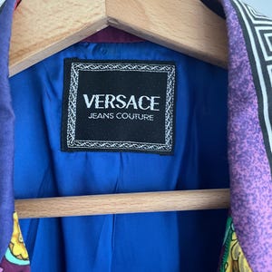 May include: A Versace Jeans Couture blazer with a vibrant patterned design in purple, black, and white. The interior lining is a bright blue, and the label is visible. The blazer is hanging on a wooden hanger.