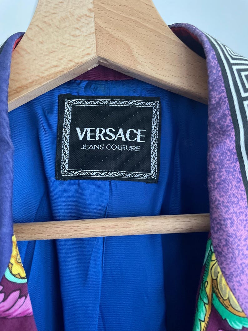 May include: A Versace Jeans Couture blazer with a vibrant patterned design in purple, black, and white. The interior lining is a bright blue, and the label is visible. The blazer is hanging on a wooden hanger.