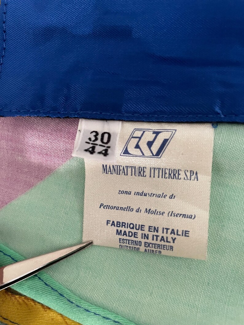 May include: A close-up of a garment tag with the text "MANIFATTURE ITTIERRE S.P.A." and "FABRIQUE EN ITALIE MADE IN ITALY". The tag also shows the size "30/44". The garment has a colorful patchwork design.