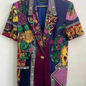 May include: A short-sleeved blazer with a vibrant floral and geometric pattern. The jacket features a sunflower, purple flowers, and gold accents. It has a blue lining and a gold button closure. The Versace label is visible.