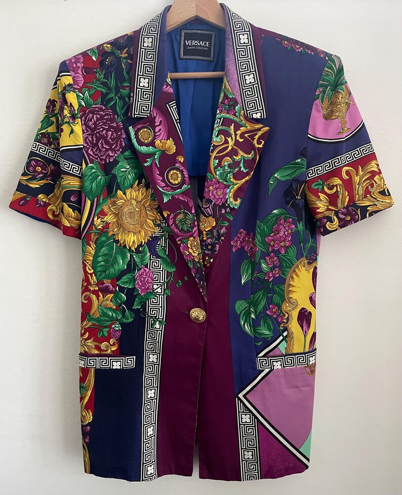 May include: A short-sleeved blazer with a vibrant floral and geometric pattern. The jacket features a sunflower, purple flowers, and gold accents. It has a blue lining and a gold button closure. The Versace label is visible.