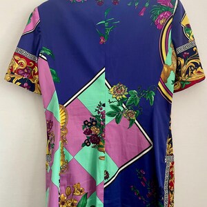 May include: A short-sleeved blazer with a vibrant floral and geometric pattern. The design incorporates shades of blue, pink, green, and gold, with ornate detailing and a Greek key border. The blazer has a collared neckline.