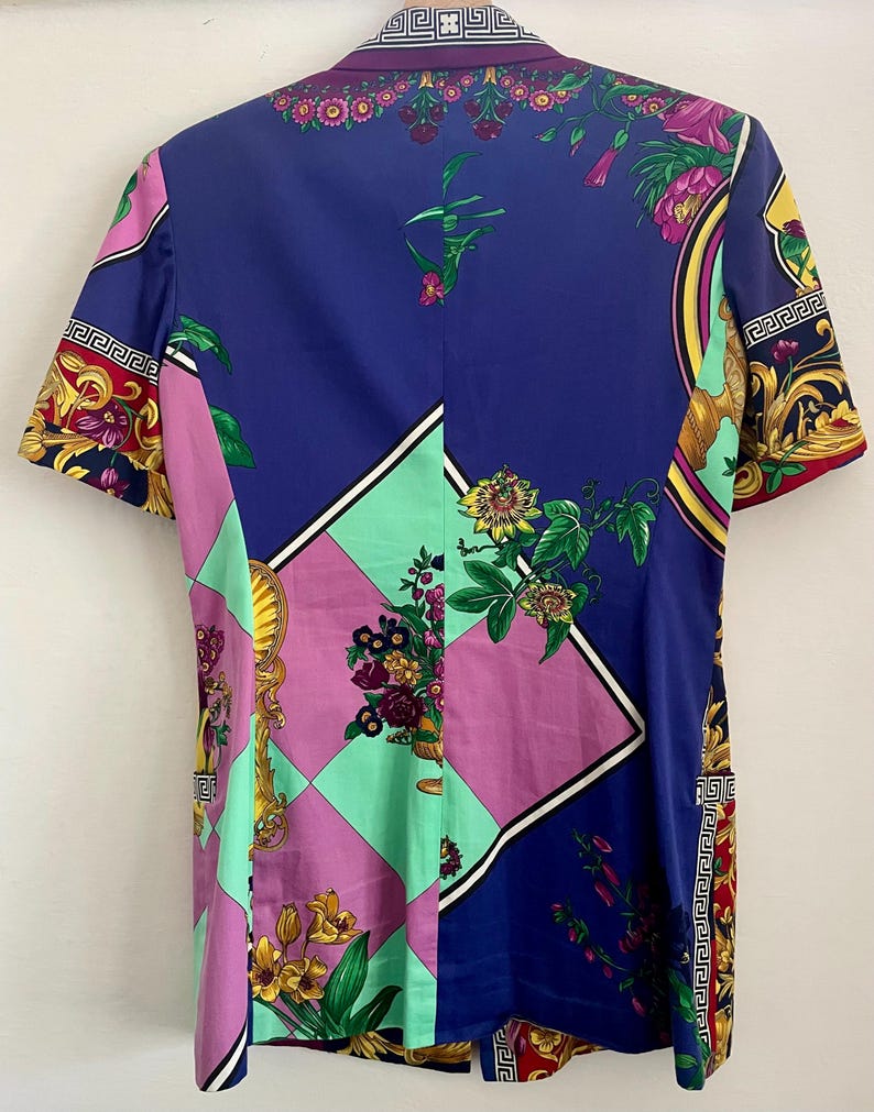 May include: A short-sleeved blazer with a vibrant floral and geometric pattern. The design incorporates shades of blue, pink, green, and gold, with ornate detailing and a Greek key border. The blazer has a collared neckline.