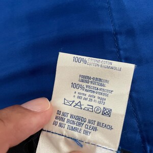 May include: A close-up of a clothing tag with text in multiple languages. The tag is white with black text and includes washing instructions. The tag is against a blue fabric.