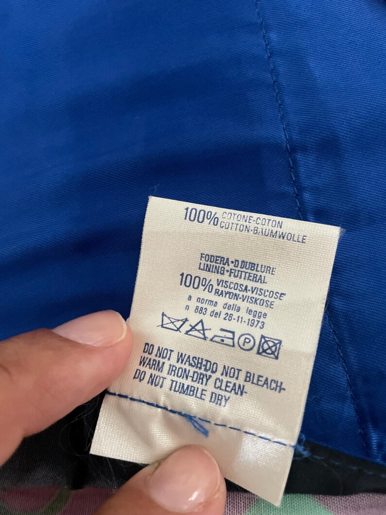 May include: A close-up of a clothing tag with text in multiple languages. The tag is white with black text and includes washing instructions. The tag is against a blue fabric.