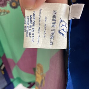 May include: A close-up of a tag from a garment, with text that reads "MANIFATTURE ITTIERRE SPA" and "FABRIQUE EN ITALIE MADE IN ITALY". The tag is white with blue and black text. The background shows a colorful floral pattern.
