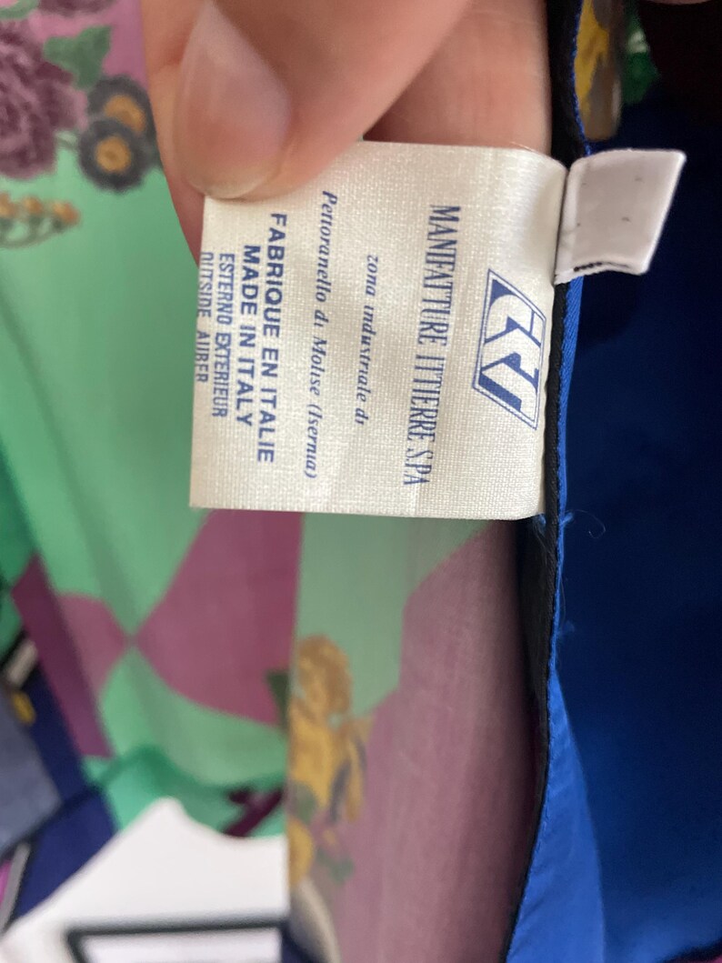 May include: A close-up of a tag from a garment, with text that reads "MANIFATTURE ITTIERRE SPA" and "FABRIQUE EN ITALIE MADE IN ITALY". The tag is white with blue and black text. The background shows a colorful floral pattern.