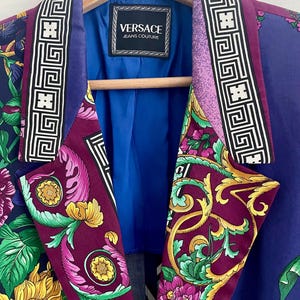 May include: A vibrant purple blazer with a floral pattern, featuring yellow sunflowers and pink flowers. The lapels have a geometric design in black and white. The label reads "VERSACE JEANS COUTURE". The blazer is hanging on a wooden hanger.