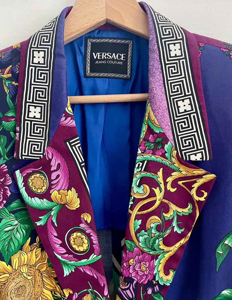 May include: A vibrant purple blazer with a floral pattern, featuring yellow sunflowers and pink flowers. The lapels have a geometric design in black and white. The label reads "VERSACE JEANS COUTURE". The blazer is hanging on a wooden hanger.