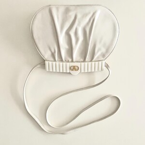 Pode incluir: Uma bolsa de m&atilde;o de couro branco com uma parte superior arredondada e franzida e uma al&ccedil;a de ombro branca. A bolsa tem um fecho dourado com um design de c&iacute;rculo duplo.
