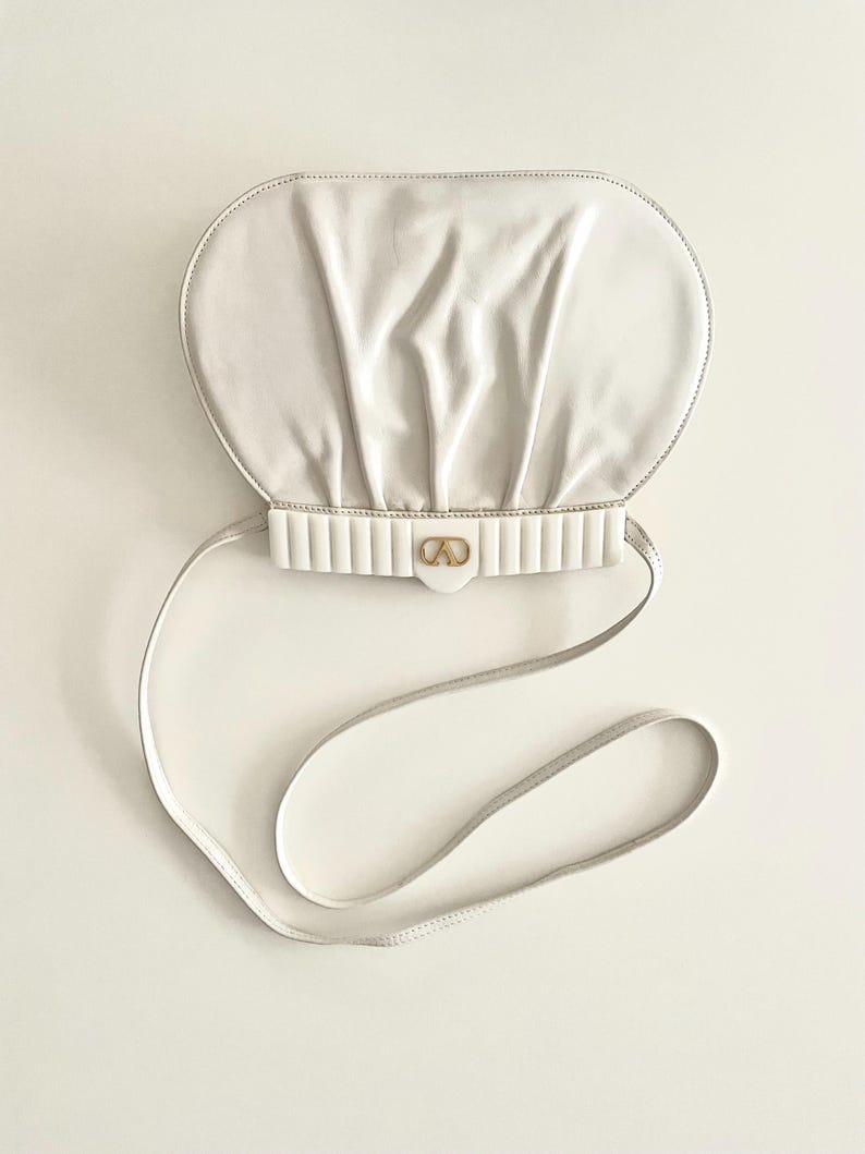 Pode incluir: Uma bolsa de m&atilde;o de couro branco com uma parte superior arredondada e franzida e uma al&ccedil;a de ombro branca. A bolsa tem um fecho dourado com um design de c&iacute;rculo duplo.