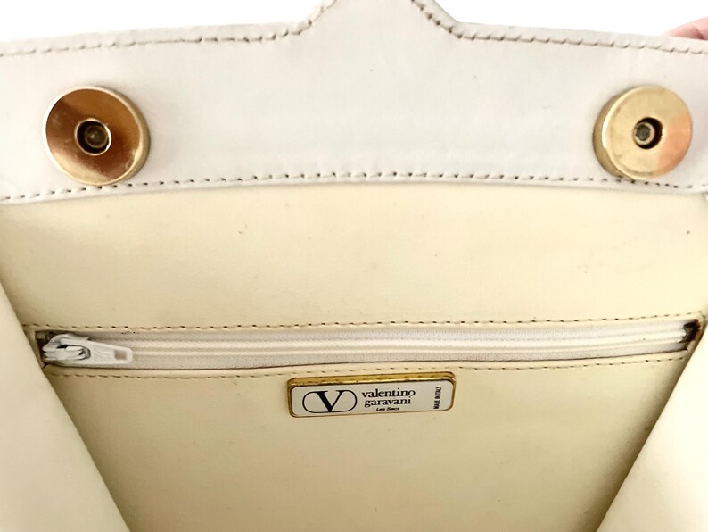 Pode incluir: O interior de uma bolsa de m&atilde;o de couro branco com um z&iacute;per dourado e uma etiqueta dourada Valentino Garavani.