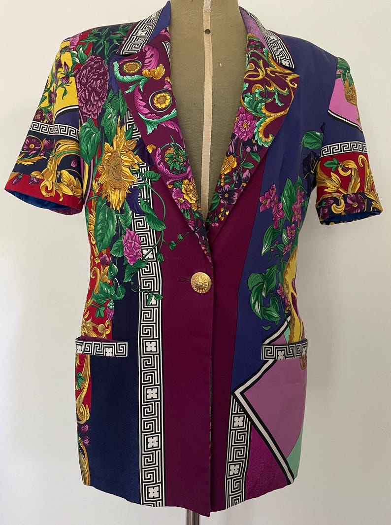 May include: A short-sleeved blazer with a vibrant floral and geometric pattern. The jacket features a mix of colors including purple, blue, yellow, and red, with a gold button closure and pocket details. The design includes sunflower and floral motifs.