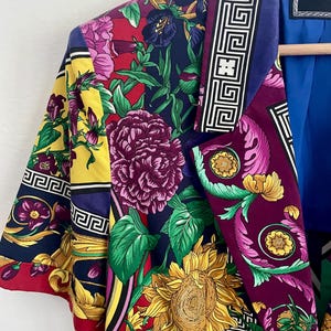 May include: A short-sleeved blazer with a vibrant floral print in shades of red, yellow, purple, and green. The blazer features a bold sunflower and other floral designs, with a geometric border along the lapel. The label reads "VERSACE JEANS CO."
