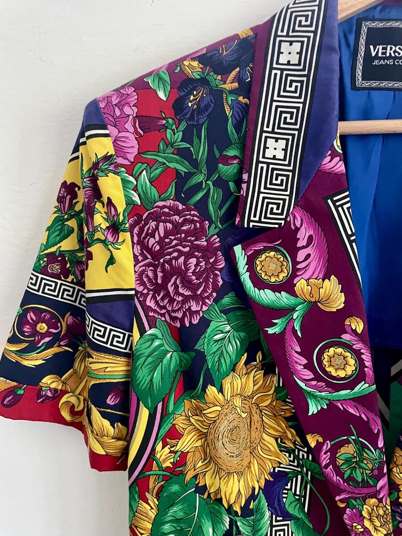 May include: A short-sleeved blazer with a vibrant floral print in shades of red, yellow, purple, and green. The blazer features a bold sunflower and other floral designs, with a geometric border along the lapel. The label reads "VERSACE JEANS CO."