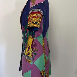 May include: A vintage blazer in a vibrant color palette of blue, purple, and green. The blazer features a bold, ornate print with gold accents and Greek key detailing. The short-sleeved jacket is displayed on a mannequin.