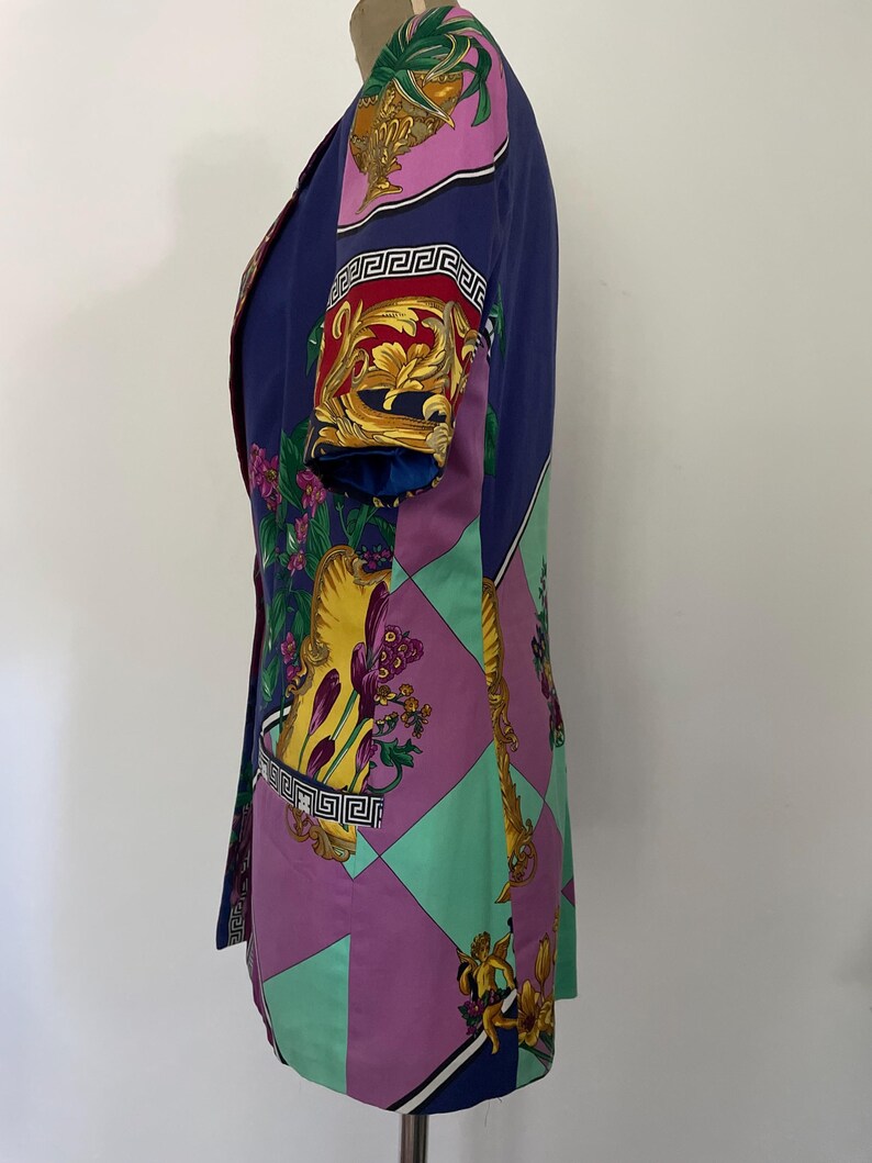 May include: A vintage blazer in a vibrant color palette of blue, purple, and green. The blazer features a bold, ornate print with gold accents and Greek key detailing. The short-sleeved jacket is displayed on a mannequin.