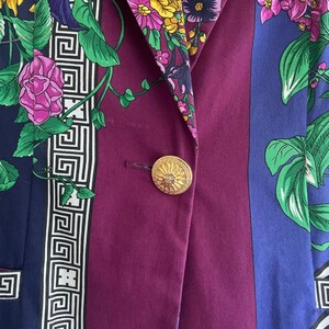 May include: A close-up of a colorful blazer with a floral pattern. The blazer features a gold button and a mix of colors, including purple, blue, and green. The floral design includes sunflowers and other flowers.