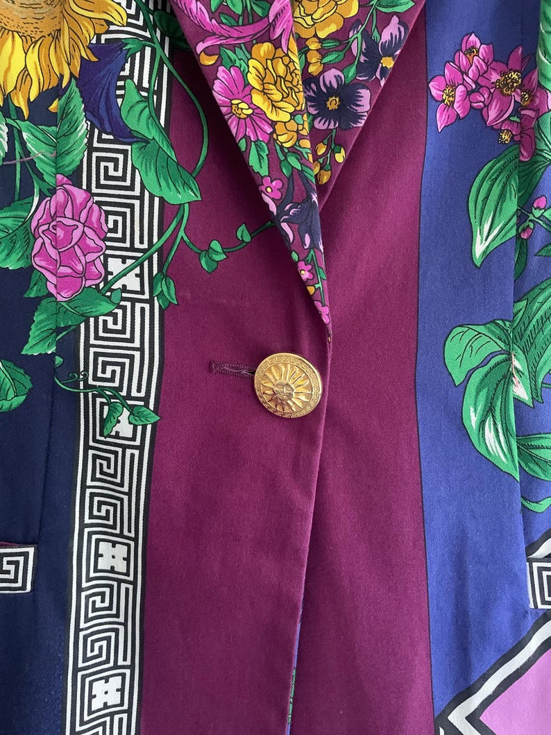 May include: A close-up of a colorful blazer with a floral pattern. The blazer features a gold button and a mix of colors, including purple, blue, and green. The floral design includes sunflowers and other flowers.