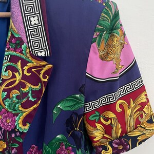 May include: A short-sleeved, patterned shirt with a vibrant floral and ornamental design. The shirt features a mix of colors, including purple, blue, pink, and gold. The design includes floral patterns, Greek key borders, and ornate details.
