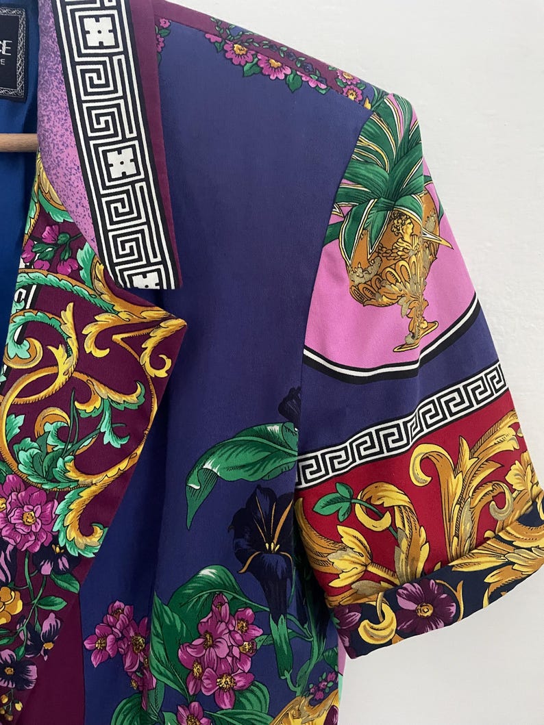 May include: A short-sleeved, patterned shirt with a vibrant floral and ornamental design. The shirt features a mix of colors, including purple, blue, pink, and gold. The design includes floral patterns, Greek key borders, and ornate details.
