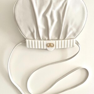 Pode incluir: Uma bolsa de m&atilde;o de couro branco com um fecho dourado e uma al&ccedil;a branca. A bolsa tem uma forma arredondada ligeiramente inchada.
