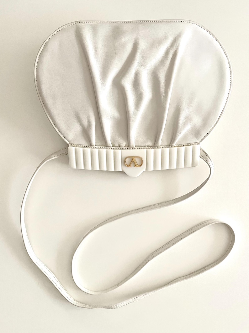 Pode incluir: Uma bolsa de m&atilde;o de couro branco com um fecho dourado e uma al&ccedil;a branca. A bolsa tem uma forma arredondada ligeiramente inchada.