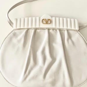 Pode incluir: Uma bolsa de m&atilde;o de couro branco com um logotipo dourado e um fecho de aba branco com nervuras. A bolsa tem uma al&ccedil;a longa branca.