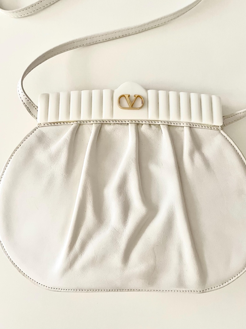 Pode incluir: Uma bolsa de m&atilde;o de couro branco com um logotipo dourado e um fecho de aba branco com nervuras. A bolsa tem uma al&ccedil;a longa branca.