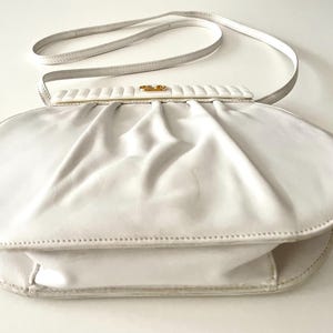 Pode incluir: Uma carteira de m&atilde;o de couro branco com um fecho dourado. A carteira tem um design plissado e uma al&ccedil;a longa.