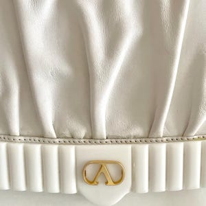 Pode incluir: Bolsa de m&atilde;o de couro branco com um logotipo dourado e uma guarni&ccedil;&atilde;o branca canelada.